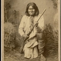 History. a old photo of a Native American kneeling pulling a rifle in the bushes.