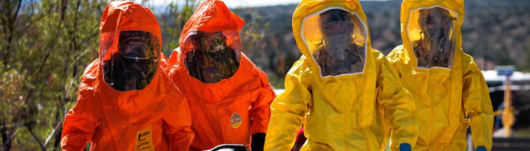 Civil Support Team. image of Four hazmat personals one wearing yellow hazmats and the other two wearing orange hazmat suits carrying stretcher. In the desert background.
