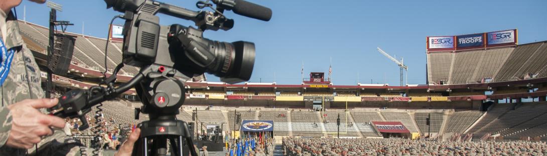 PAO banner. Military personnel is filming the celebration event, panning a video camera on a tripod that's a short black metal bar in front of him to prevent falling off the stage.