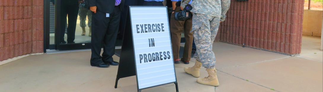 Exercise Branch Image in front of the door of the EOC, a sign says exercise in progress. People are walking in the background of the sign.