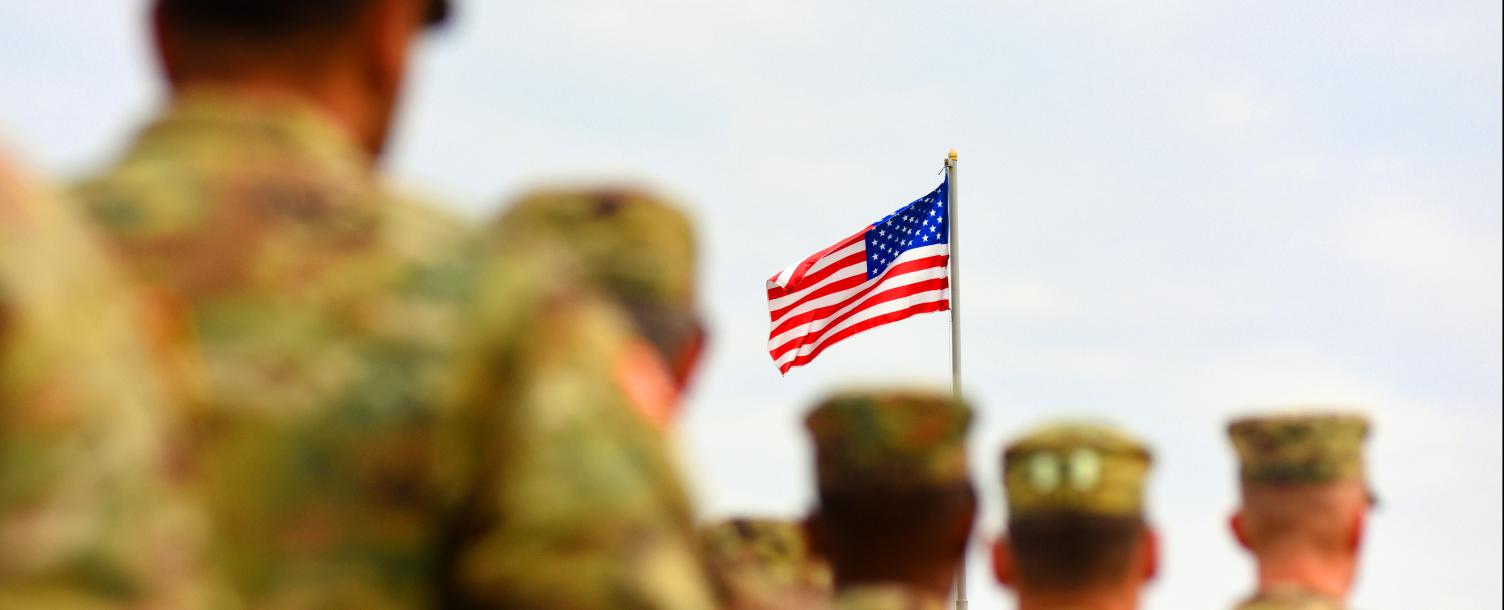 Slightly blurred backs of the military soldiers in Multicam lineup information. With the American flag in focus on the pole waiting in the air.